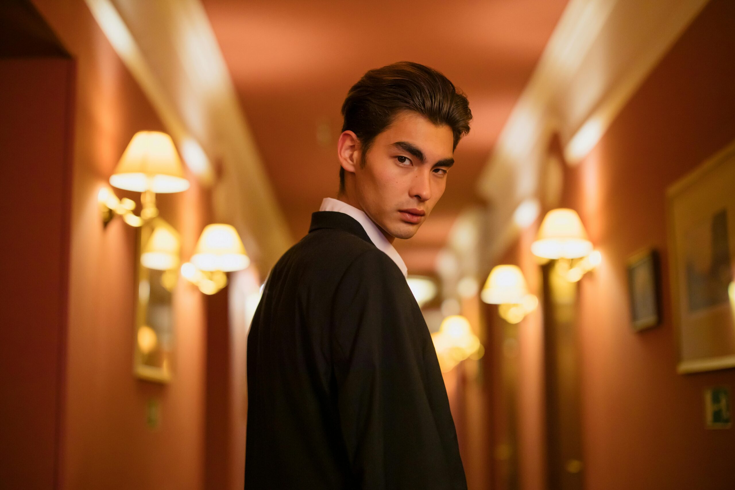 Young man in a black suit standing confidently in a warmly lit hotel corridor.